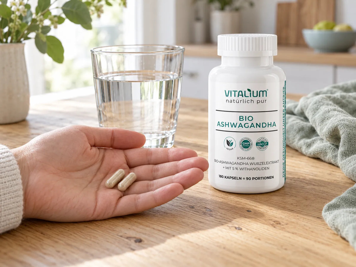 Hand mit zwei Ashwagandha-Kapseln, daneben ein Glas Wasser und die Dose auf einem Holztisch